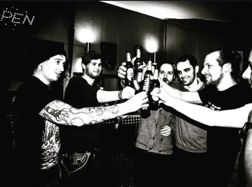 Sechs Männer stoßen mit Flaschen an, der Raum ist schwach beleuchtet.