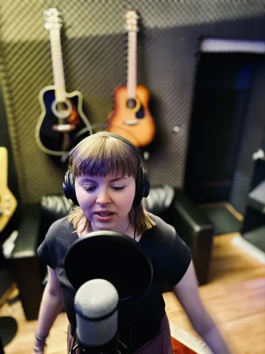 Frau singt im Aufnahmestudio, Mikrofon vor ihr, Akustikgitarren im Hintergrund.