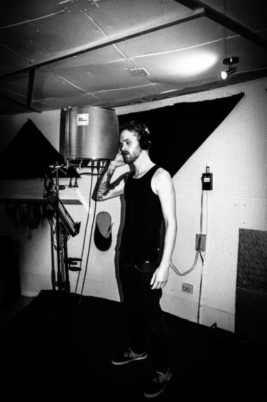 Ein Mann steht im Studio und spricht ins Mikrofon, im Hintergrund technische Geräte.