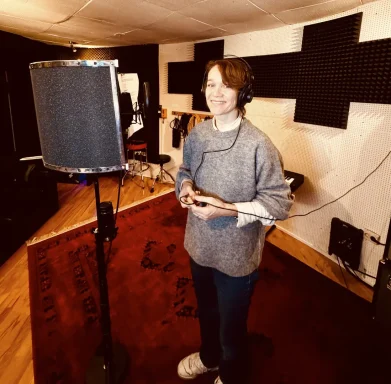 Person steht in einem Tonstudio nahe einem Mikrofon und trägt Kopfhörer.