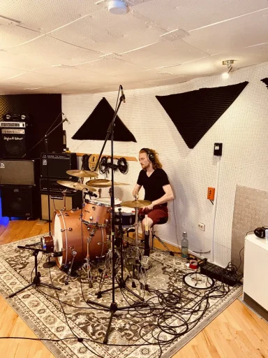 Ein Schlagzeuger sitzt in einem Studio und spielt Schlagzeug. Akustische Elemente sichtbar.