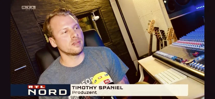 Mann sitzt an einem Tisch mit Mischpult, im Hintergrund Musikgeräte und dunkle Wände.