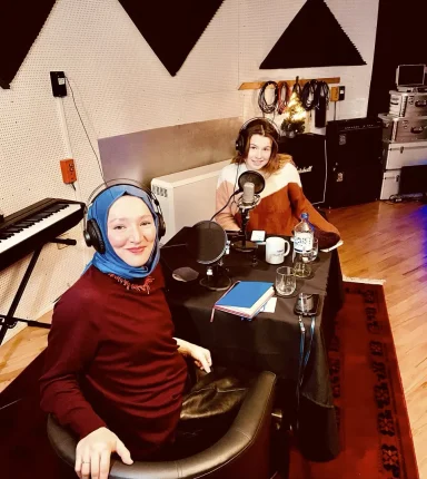 Zwei Frauen im Gespräch in einem Podcast-Studio, Mikros und Notizen sichtbar.