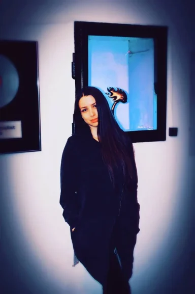 Eine Person mit langen, dunklen Haaren steht vor einem blauen Hintergrund und Bilderrahmen.