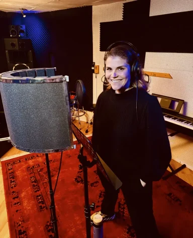 Lächelnde Person im Studio, trägt Kopfhörer und steht am Mikrofon.