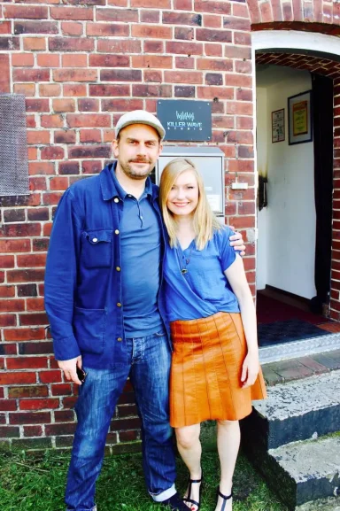 Zwei Personen stehen lächelnd vor einer Backsteinmauer, eine mit Cap, die andere in kurzer Bluse.