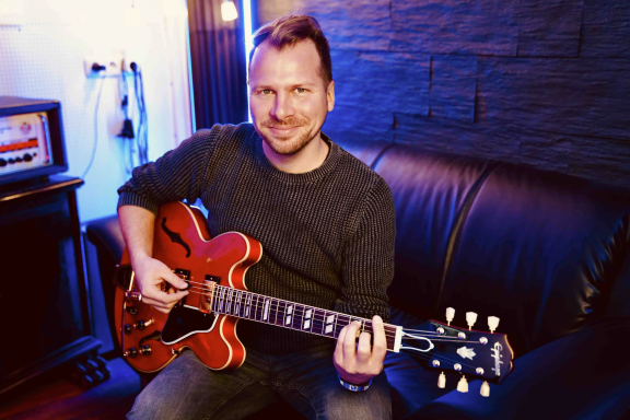 Ein lächelnder Mann sitzt mit einer roten E-Gitarre auf einem Sofa.