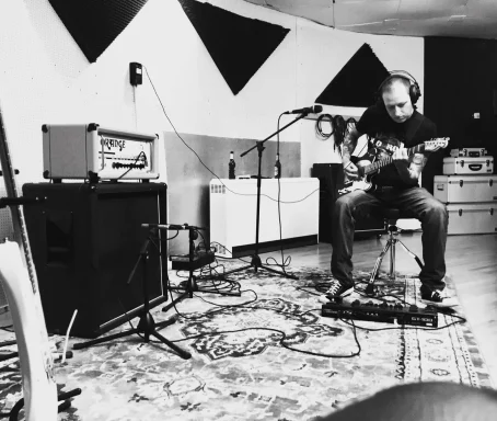 Musiker sitzt im Aufnahmestudio und spielt Gitarre. Ausstattung im Hintergrund sichtbar.