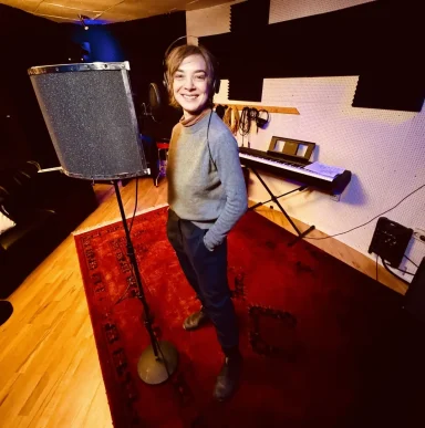 Junge steht in einem Tonstudio neben einem Mikrofon, lächelt freundlich.