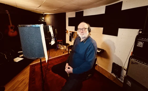 Ein älterer Mann sitzt in einem Tonstudio vor einem Mikrofon, umgeben von Instrumenten.