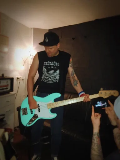 Mann spielt auf einem hellblauen Bass, trägt ein schwarzes ärmelloses Shirt und eine Cap.