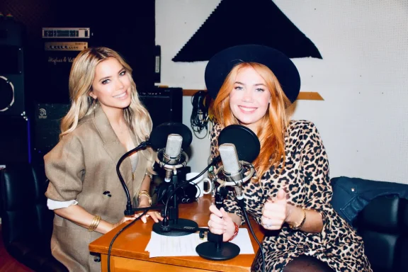 Zwei Frauen stehen vor Mikrofonen in einem Studio, lächeln und posieren freundlich.