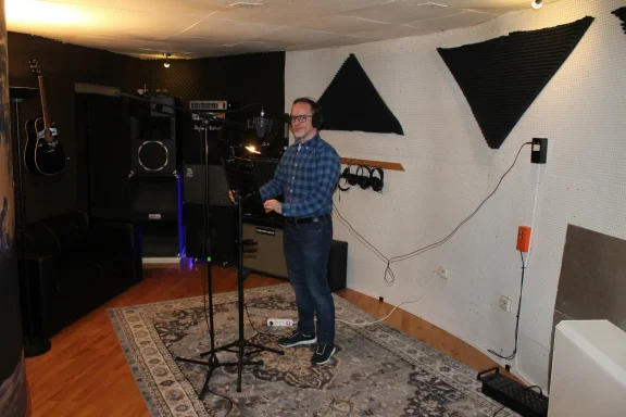 Mann in einem Tonstudio steht vor einem Mikrofon, umgeben von Musikequipment.