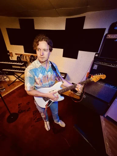 Mann spielt E-Gitarre in einem Studio mit Musikgeräten im Hintergrund.