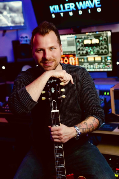 Mann mit Gitarre in einem Tonstudio, umgeben von Musik-Equipment.