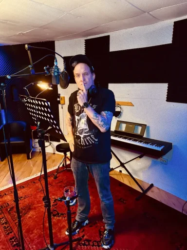 Mann steht im Studio am Mikrofon, umgeben von Musikinstrumenten.