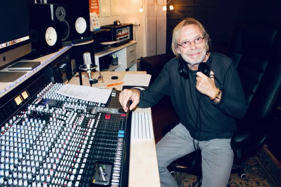 Mann in einem Tonstudio, lächelnd am Mischpult mit Kopfhörern um den Hals.