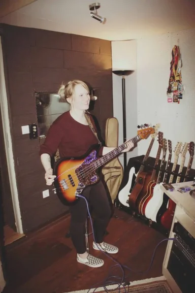 Frau spielt E-Bass in einem Musikraum mit mehreren Gitarren im Hintergrund.