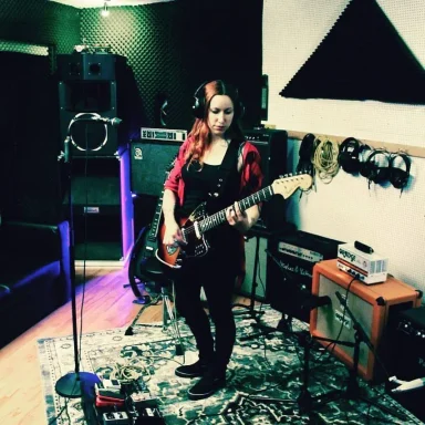 Frau mit rotem Haar spielt E-Gitarre in einem Proberaum. Kabel und Lautsprecher sichtbar.