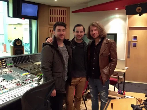 Drei Männer in einem Tonstudio, lächelnd vor einem Mischpult.