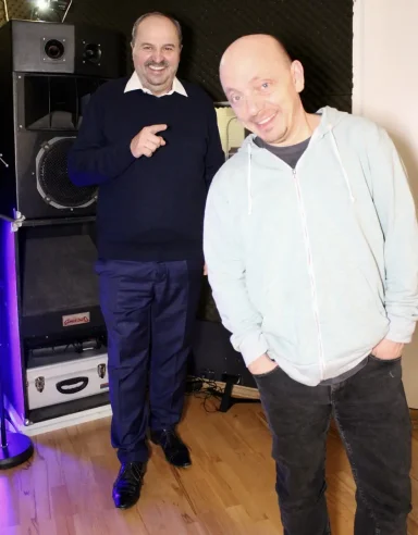 Zwei Männer stehen in einem Raum, einer lächelt und der andere zeigt mit dem Finger.