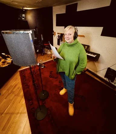 Frau in einem Aufnahmestudio, spricht ins Mikrofon, trägt grüne Kleidung und gelbe Schuhe.