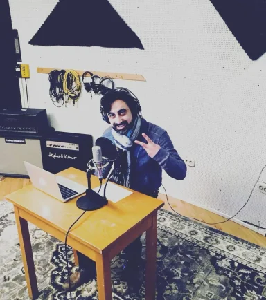 Mann mit Brille und Schal sitzt an einem Tisch mit Laptop und Mikrofon in einem Studio.