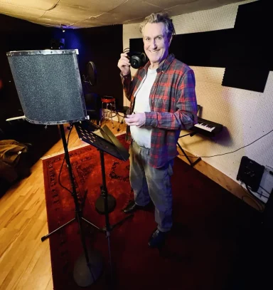 Ein Mann steht in einem Tonstudio neben einem Mikrofon und hält ein Aufnahmegerät.