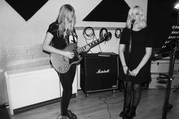 Zwei Musikerinnen in einem Studio, eine spielt Gitarre, die andere singt.