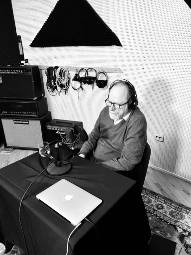 Ein Mann in einem Sessel trägt Kopfhörer und sitzt vor einem Tisch mit Mikrofon und Laptop.