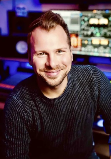 Lächelnder Mann in einem Studio mit Musik-Equipment im Hintergrund.