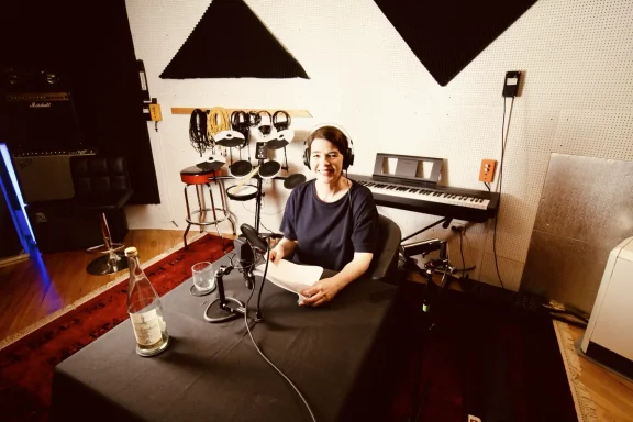 Frau sitzt an einem Tisch im Studio mit Mikrofon, Klavier und Musikinstrumenten im Hintergrund.